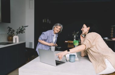 Cep telefonuyla konuşurken dizüstü bilgisayarı işaret eden olumlu bir genç kız ve yemek hazırlayan ve ekranda dikkatle bakan bir anne.