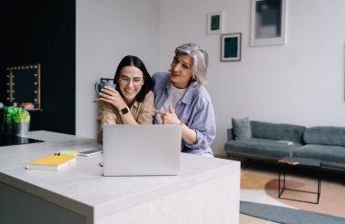 Yaşlı anne bilgisayardaki video konuşmasında eğlenirken, neşeli genç bayan bir kupa sıcak içecekle gülüyor.