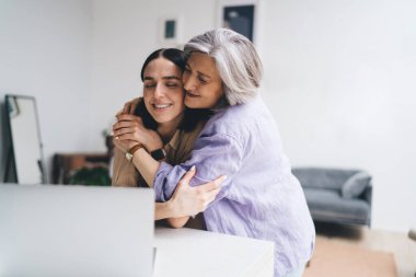 Gündelik kıyafetlerle birbirini seven anne ve kız evdeki ışık minimalist odada dizüstü bilgisayarla otururken birbirlerine sarılıyorlar.
