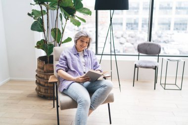 Yüksek açılı, günlük kıyafetli, yeşil bitki okuma romanının yanındaki rahat koltukta dinlenen, huzurlu bir atmosferde yaşlı bir kadın.