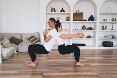 Koltukta oturan genç bir kadının vücut manzarası Utkatasana 'ya poz verirken evdeki rahat oturma odasında antrenörün sırtında ağırlığı dengeliyor.