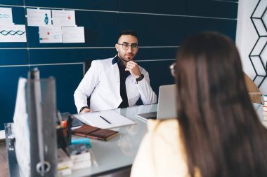 Ciddi bir erkek doktor, doktor muayenehanesinde randevusu sırasında el pençe divan duran ve ekinlere bakan isimsiz bir hasta.
