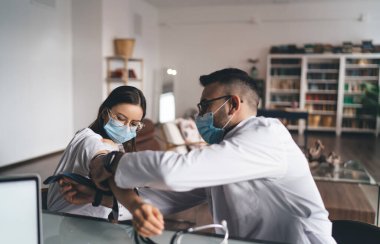 Kan basıncını ölçen koruyucu maskeli genç bir erkek doktorun yan görüntüsü. Kadın hasta klinikte dizüstü bilgisayarla masada oturuyor.