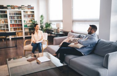 Yastıklı koltukta oturan ve modern ofisteki profesyonel psikoterapistle sorunları paylaşan sakallı erkek müşterinin yan görüntüsü.