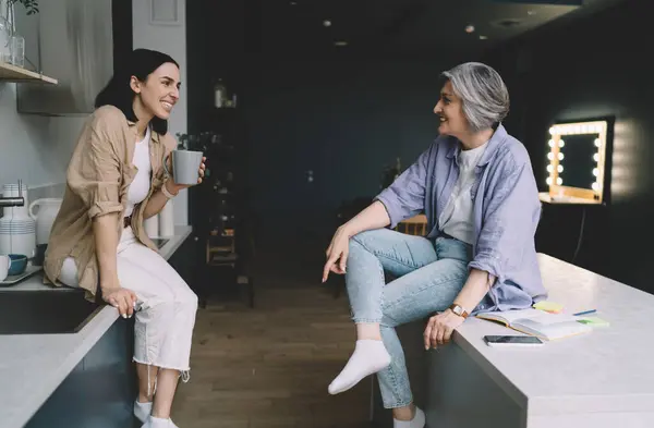 stock image Side view of young woman sitting on kitchen worktop having hot drink while talking to elderly mother sitting on countertop with legs crossed