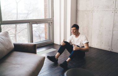 Aktif kıyafetli neşeli bir adam büyük pencerenin yanında oturmuş spordan keyif alıyor ve gülümsüyor.