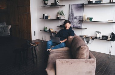 Temiz tıraşlı bir erkek çalışanın kameraya bakıp rahat bir koltukta oturup evdeki uzak projede dizüstü bilgisayar üzerinde çalışmasının yan görüntüsü