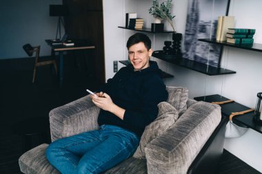 Elinde cep telefonuyla gezen ve hafta sonu boş zamanlarında oturma odasındaki rahat koltukta rahatlarken gülümseyen ve yüzünü çeviren olumlu genç adam.