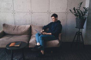 Yüksek odaklı erkek girişimci, internet üzerinden iş projesi üzerinde çalışırken internet üzerinden kitap yazma bilgisiyle sehpanın yanındaki koltukta oturuyor.