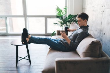 Akıllı telefon ekranına bakan ve evdeki rahat oturma odasındaki kanepede dinlenirken mesajları arayan genç adamın yan görüntüsü.