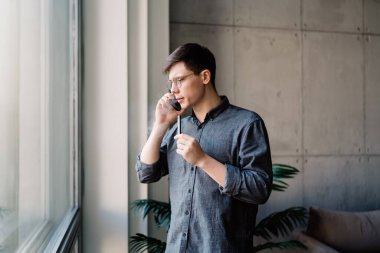 Sakin erkek, günlük kıyafetler ve gözlük takıyor. Elinde kalemle pencerenin yanında dikilip gözlerini kaçırırken cep telefonuyla konuşuyor.