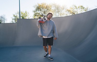 Gri kapüşonlu kaykaycı vücudunun tamamı kaykay parkındaki bowling rampasında dinlenirken uzun tahta taşıyor.