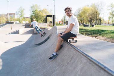 Paten parkındaki eğitimde kaykay yaparken dinlenen ve kentsel çevreye karşı kameraya bakan düşünceli erkek kaykaycının yan görüntüsü