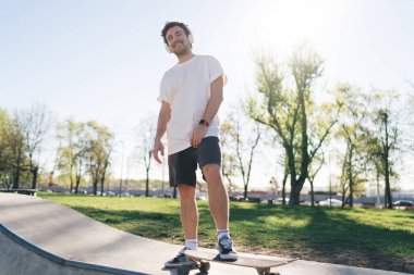 Kulaklıklardan müzik dinlerken, kaykayın üstünde tek bacakla rampanın üzerinde dururken kameraya bakan neşeli, genç bir erkek patencinin alçak açısı.