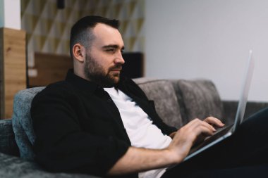 Yoğunlaştırılmış erkek girişimcinin yan görünüşü kanepeye oturup dizüstü bilgisayarında yazı yazması modern çalışma alanında online iş projesinde sıkı çalışması
