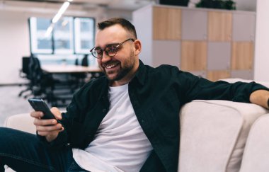 Modern çalışma ortamında sakallı, rahat bir koltukta oturup, akıllı telefondan sohbet eden sosyal ağda çalışan pozitif girişimci.