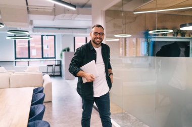 Kameraya bakarak şirkette çalışırken cam duvarın yanındaki çağdaş çalışma alanında yürüyen pozitif sakallı bir girişimci.