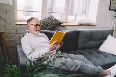 Hafif hizmetçi kıyafetli yaşlı beyefendi rahat bir kanepeye uzanıyor ve evdeki oturma odasında ilginç bir kitap okuyor.