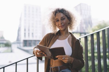 Şehirli binaların bulanık arka planında dururken günlük kıyafetleriyle gülümseyen kıvırcık saçlı pozitif etnik kız öğrenci.