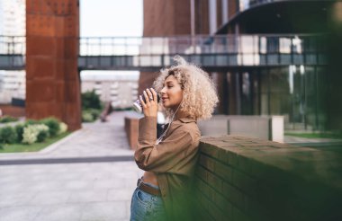 Kenar manzaralı, kulaklıklı, şehir sokaklarında dikilmiş, kağıt bardaktan kahve içen sarışın, Afro-Amerikalı genç bir kadın.