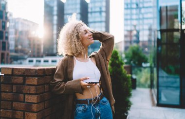 Sokak tarzı giyinmiş kıvırcık sarı saçlı pozitif genç bir kadın. Tuğla çitin yanında duruyor ve saçlarına dokunuyor. Şehir merkezinde sabah vaktinin keyfini çıkarırken müzik dinliyor.