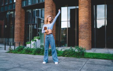 Tam boy etnik bayan öğrenci günlük kıyafetler giyiyor üniversite binasının yanında elinde kitaplarla ayakta duruyor ve kollarını kaldırarak sevinçlerini ifade ediyorlar.