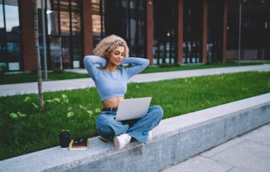 Genç Afrikalı Amerikalı serbest çalışan kadın kahve fincanı ve not defteriyle bacak bacak üstüne atarak ellerini boynunun arkasına koyuyor ve uzaktan bilgisayarın ekranına bakıyor.