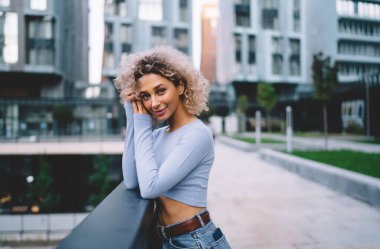 Sarı kıvırcık saçlı, mavi günlük kıyafetler giyen terasa yaslanmış ve kameraya bakan Afro-Amerikan kadın.