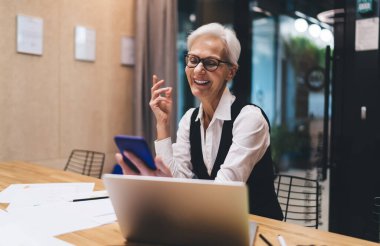 Neşeli, gri saçlı, resmi kıyafetli, cep telefonuyla video konuşması yapan, iş yerinde dizüstü bilgisayarlı ve belgeli yaşlı bayan çalışanı.