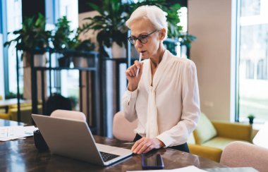 Kalemle dudakları birbirine değdirip iş projesi üzerinde çalışırken ekranda dizüstü bilgisayara bakan odaklanmış yaşlı bir kadın.