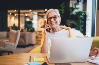 Gözlüklü pozitif yaşlı bir iş kadını bilgisayarla masaya otururken kameraya bakıyor ve cep telefonuyla konuşuyor.