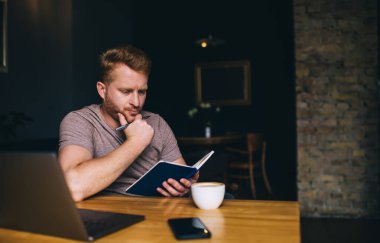 Düşünceli, sakallı, günlük giysiler içinde, elinde defter ve kalemle evden bilgi analiz edip netbook ile uzaktan eğitim alan bir erkek öğrenci.