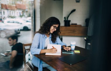 Kafeteryada dizüstü bilgisayar ve kağıt kahve fincanı ile masada otururken, günlük giysiler ve gözlüklerle not defteri ve cep telefonuna not defteri yazıp gezinen memnun bir kadın.