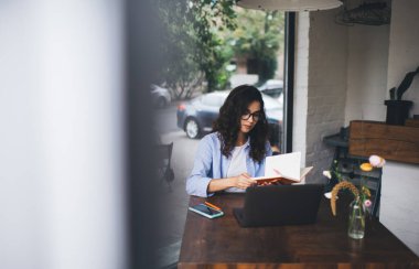 Kafede internet üzerinde çalışırken günlük kıyafet ve gözlük takan konsantre kadın girişimci planlamacıda notlar okuyor.
