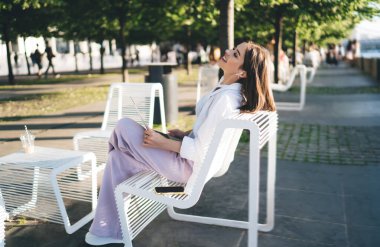 Gündüz kıyafetleriyle bankta oturan, akıllı telefonlu ve güneşli bir günde parkta dinlenen genç bir kadının yan görüntüsü.