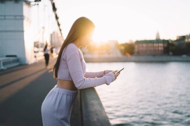 Mor kıyafetli zayıf bir bayanın dalgalı suyun yanında duran setin kenarına bakması ve gün batımında cep telefonuyla mesaj göndermesi.