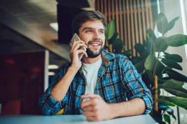 Yeşil saksı bitkisinin yanındaki masada oturan ve kafeye bakarken cep telefonuyla konuşan, gündelik kıyafetli, pozitif sakallı bir erkek.