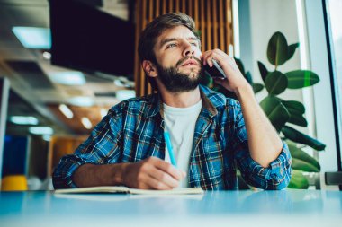Düşük açılı sakallı erkek serbest çalışan. Masada defter başında oturmuş cep telefonuyla konuşurken kalemle not alıyor.