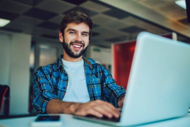 Düşük açılı pozitif sakallı erkek serbest çalışan gülümseyen ve kameraya bakan modern restoranda uzaktan kumandalı bir proje üzerinde çalışırken dizüstü bilgisayarlı ve cep telefonlu.