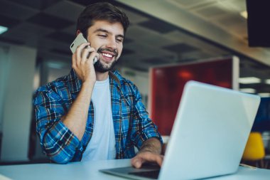 Düşük açılı pozitif sakallı serbest çalışan erkek bilgisayar başında oturmuş cep telefonuyla konuşurken ekrana bakıyor ve kafede uzaktan kumandalı yazılım şirketi için çalışıyor.
