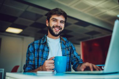 Düşük açılı pozitif sakallı erkek serbest çalışan, gülümseyen ve kameraya bakan, elinde akıllı telefon ve kahveyle masada otururken, kafede yeni başlayanlar için dizüstü bilgisayar üzerinde çalışan.