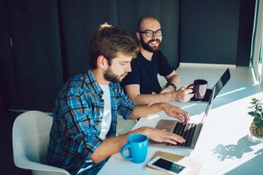 Gülümseyen sakallı serbest çalışan, şık giyinen, kahve içip bilgisayarını karıştıran, modern ofisteki iş arkadaşının yanında oturan.