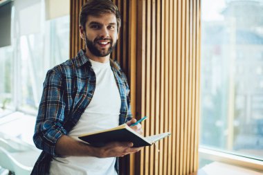 Neşeli bir erkek çalışanın kameraya bakıp gülümsemesi ve not defterine notlar yazması ve modern bir iş yerinde çalışması.