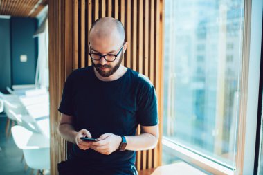 Yoğunlaşmış sakallı erkek akıllı saat takıyor cam pencerenin yanındaki masada oturuyor ve modern kafede gündüz ekrana bakarken cep telefonunu karıştırıyor.