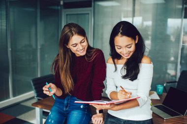 Uzun siyah saçlı gülümseyen iş arkadaşları, modern çalışma ortamında dizüstü bilgisayarla masaya yaslanırken günlük kıyafetlerini giyerler.