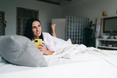 İyimser genç bayan, tembel bir sabahta yatak odasında cep telefonuyla boş vakit geçirip gözlerini kaçırırken rahat bir yatakta karnının üstünde dinleniyor.