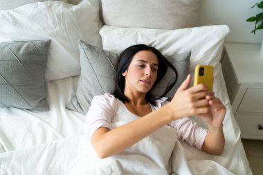 Yüksek açılı, genç, tembel bir kadın yatakta uzanıyor ve rahat yatak odasında gezinip dinlenirken akıllı telefon ekranına bakıyor.
