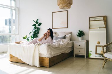 Açık pijamalı mutlu esmer kadın sabah uyandıktan sonra rahat bir yatakta uzanıyor ve cep telefonuyla konuşuyor.