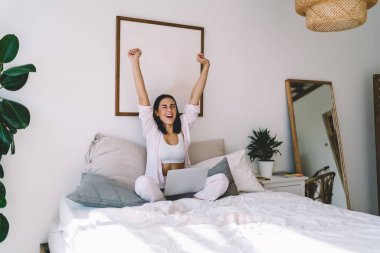 Genç, neşeli, serbest çalışan bir bayan bilgisayar ile rahat bir yatakta oturuyor gözleri kapalı ve kollarını uzatarak yeni projeyi online olarak bulmanın başarısını kutluyor.