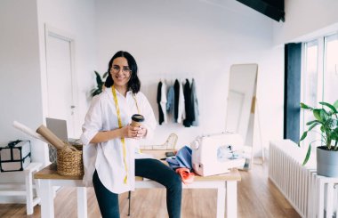 Ahşap masada oturmuş, dikiş desenleri ve sıcak kahve içen neşeli, genç, etnik kadın terzi.